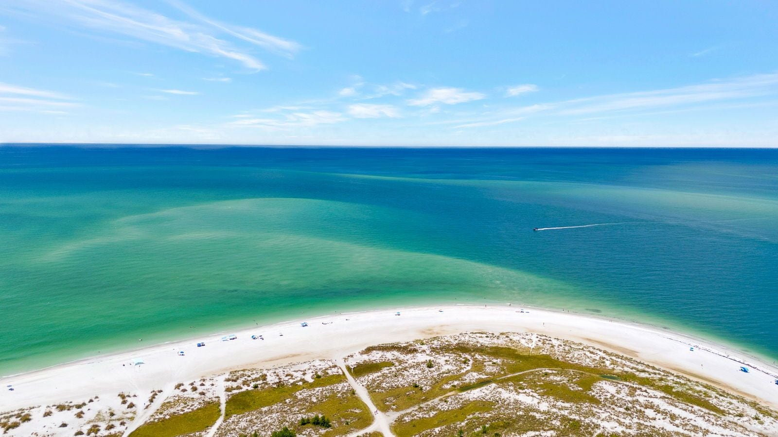 An aerial view of a beach with clear turquoise waters, a sandy shoreline, and sparse vegetation.