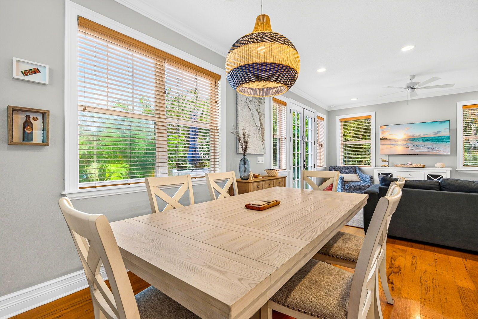 A dining area with a wooden table and chairs, a hanging light fixture, and a view of greenery through the windows.