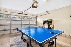 A ping pong table with a net in a room with a garage door, a table tennis table, and a table tennis machine. - Orange Crush - Private Pool Home Near Beach - By Island Real Estate (Holmes Beach)