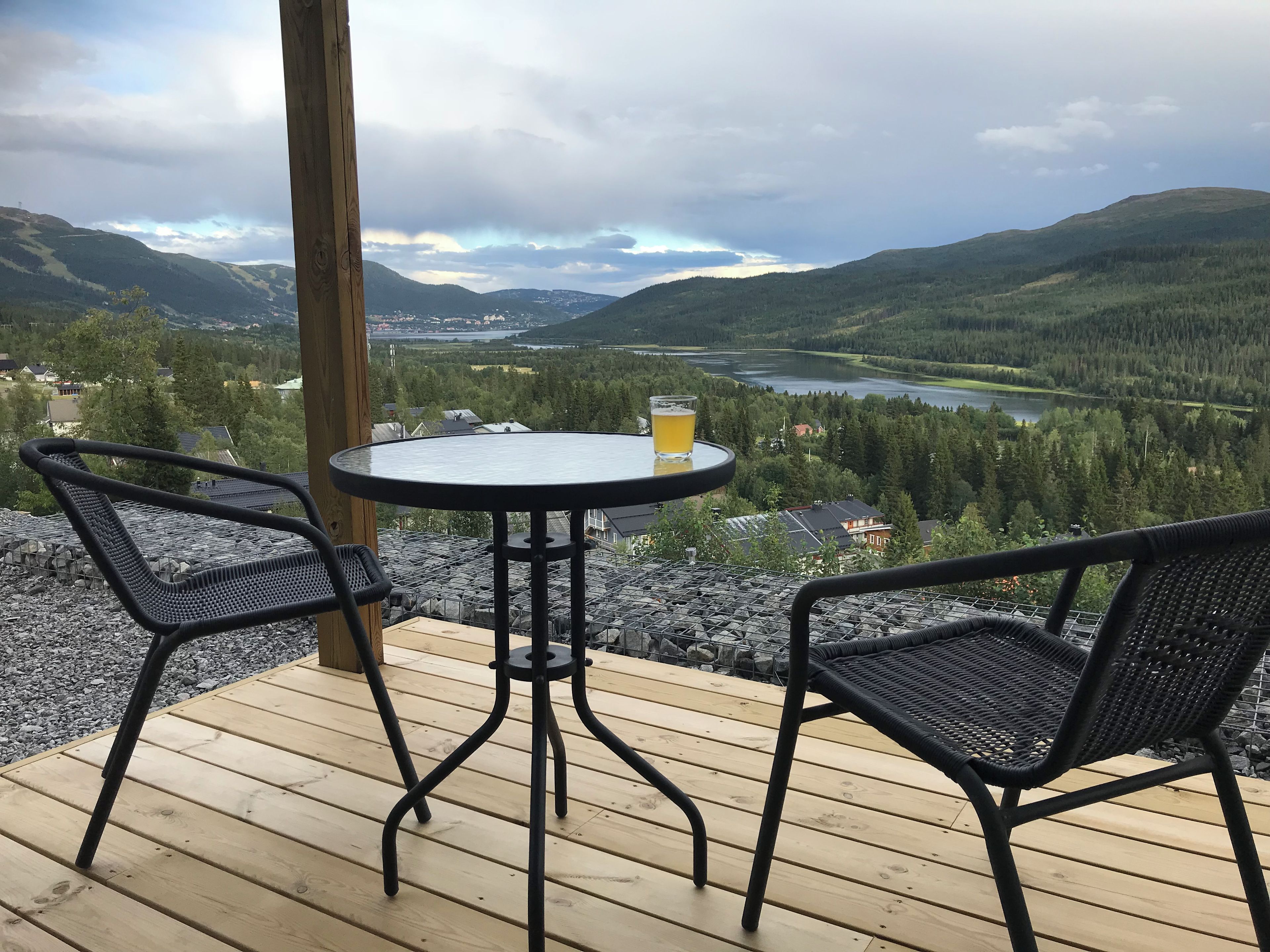 A wooden deck with a round table and two chairs, overlooking a valley with a lake and forested hills.