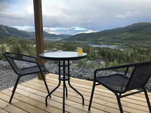 A wooden deck with a round table and two chairs, overlooking a valley with a lake and forested hills.