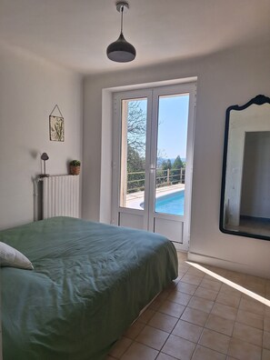 Ein Schlafzimmer mit einem Bett, einem Heizkörper, einer Topfpflanze und Blick auf ein Schwimmbad durch eine Glastür.