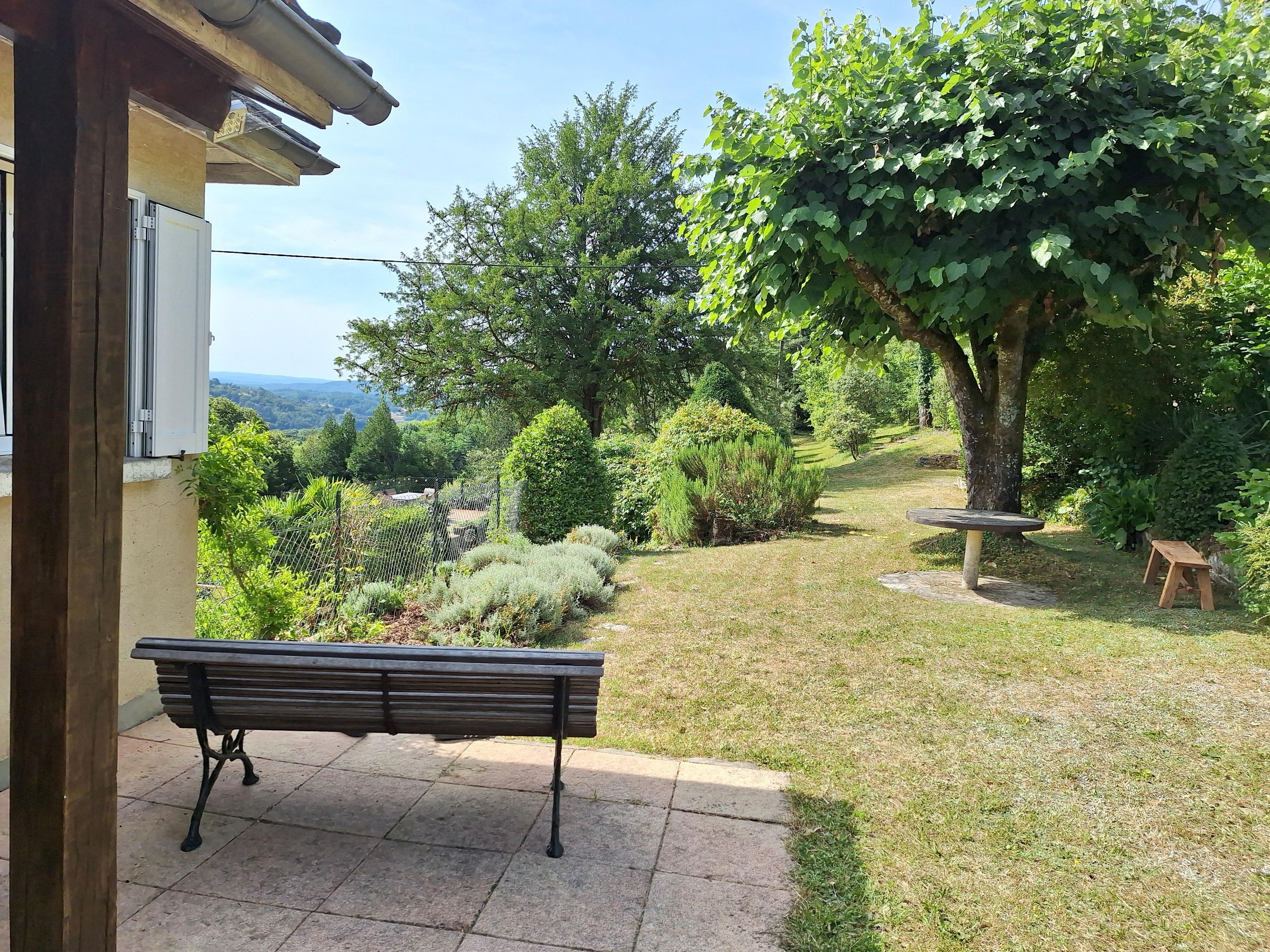 A patio with a bench, a table, and a view of a lush garden and distant hills.