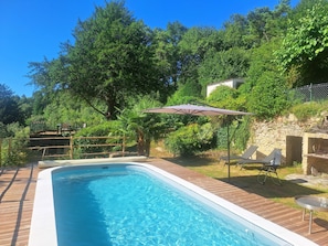 Ein Schwimmbecken mit Holzterrasse, umgeben von üppiger Vegetation und einer Steinmauer.
