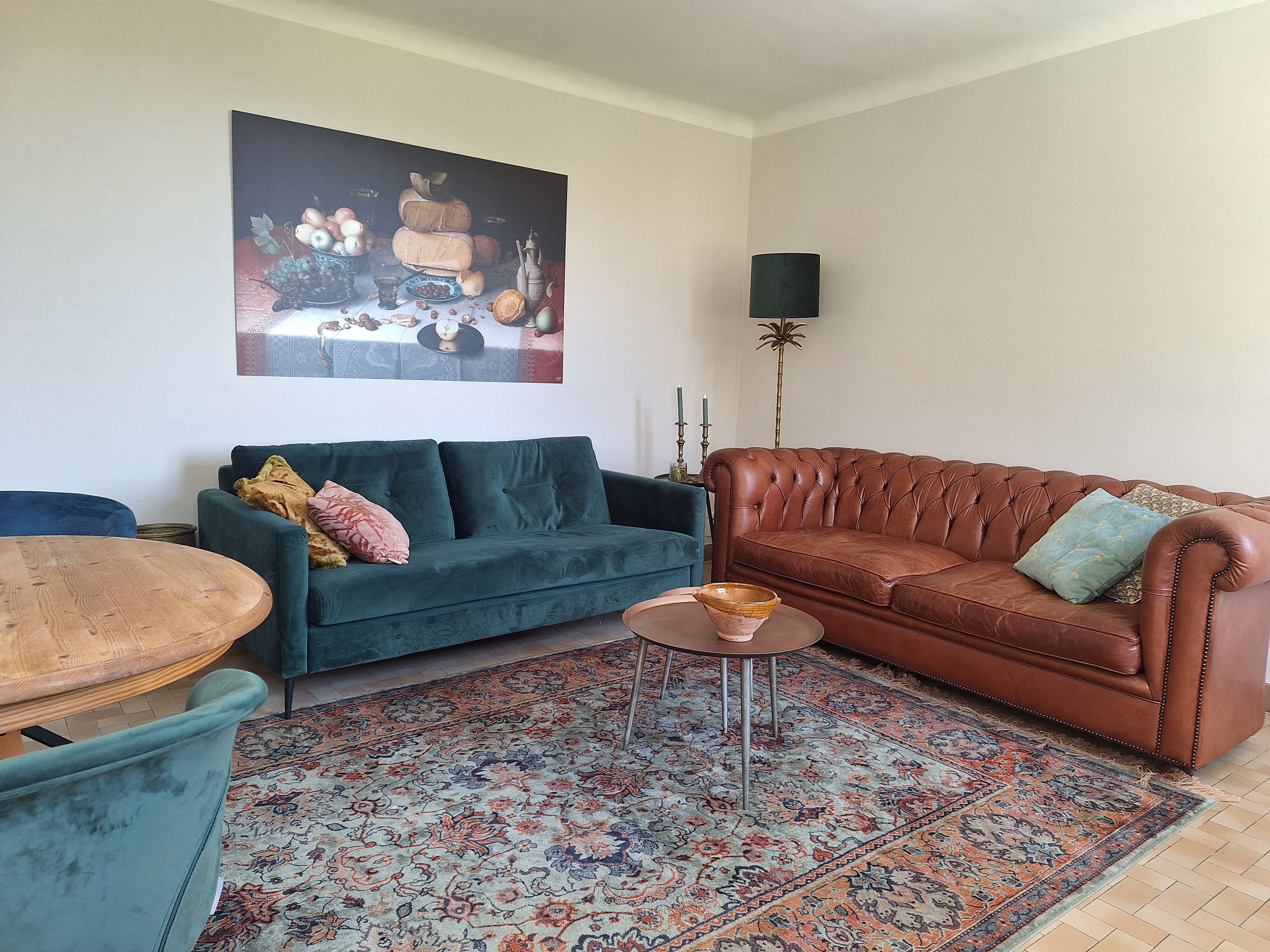 Een woonkamer met een groene bank, een bruine leren bank, een ronde houten tafel en een kamerkleed met dessin.