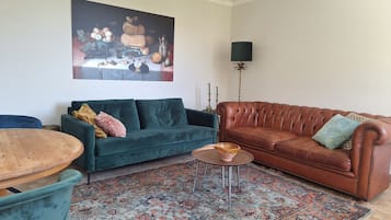 Een woonkamer met een groene bank, een bruine leren bank, een ronde houten tafel en een kamerkleed met dessin.