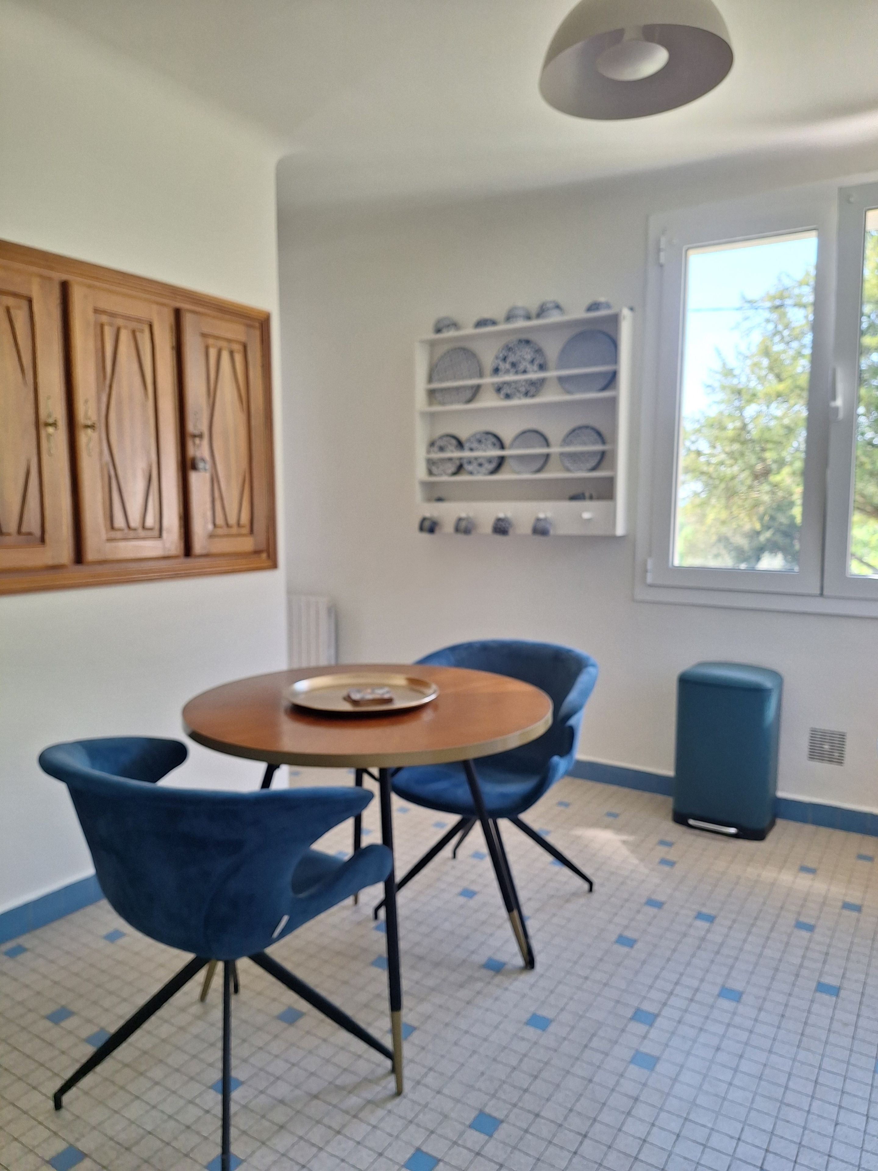 Een kamer met een ronde tafel, twee blauwe stoelen, een plank met borden, houten kasten en een raam met uitzicht.