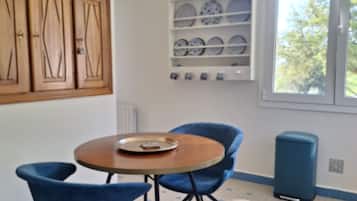 Een kamer met een ronde tafel, twee blauwe stoelen, een plank met borden, houten kasten en een raam met uitzicht.