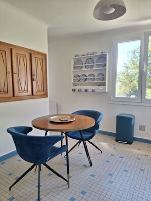 Une pièce avec une table ronde, deux chaises bleues, une étagère remplie d’assiettes, des meubles en bois et une fenêtre donnant sur un beau paysage.