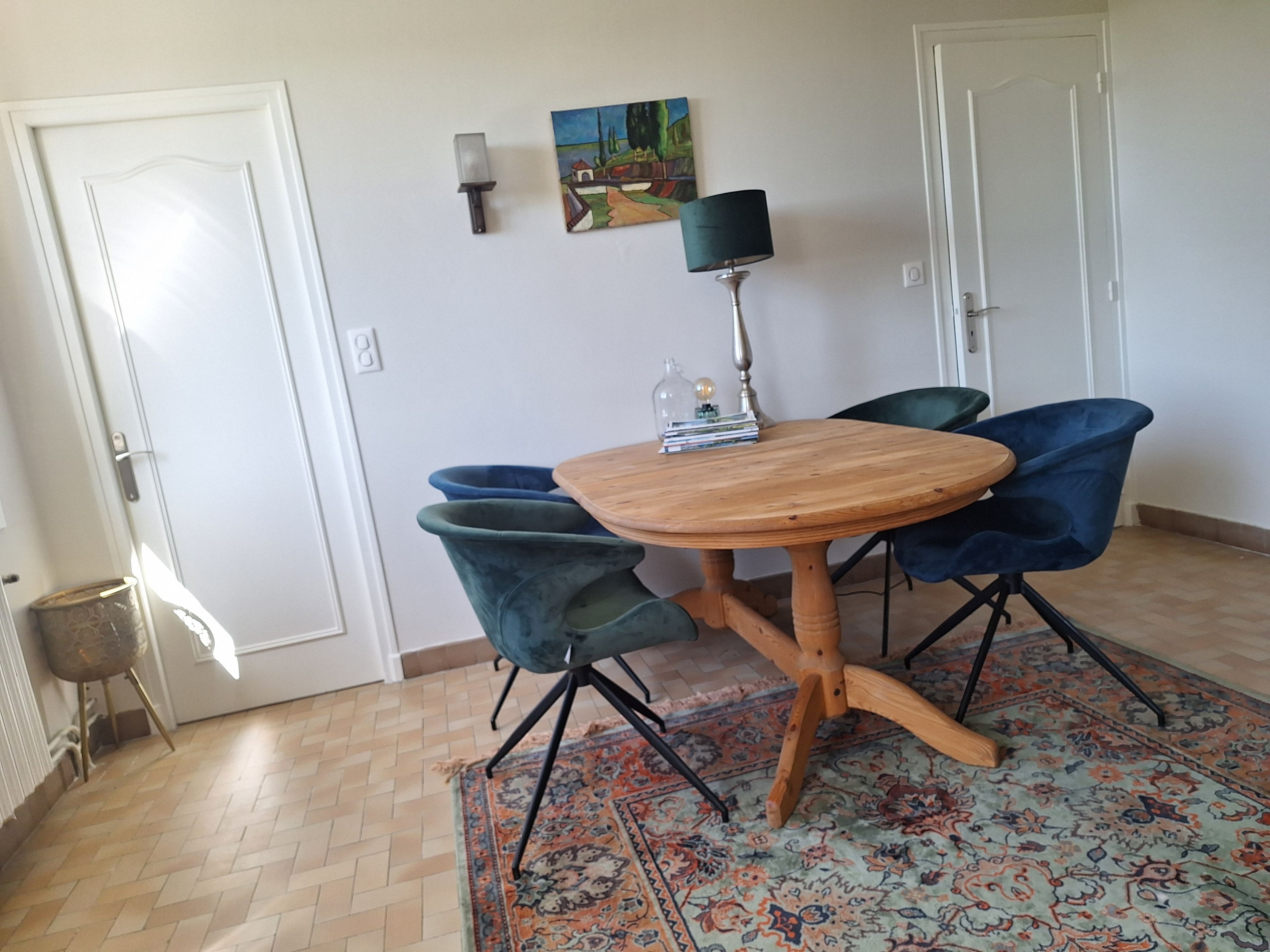 A dining area with a round wooden table, two blue chairs, a patterned rug, and a painting on the wall.