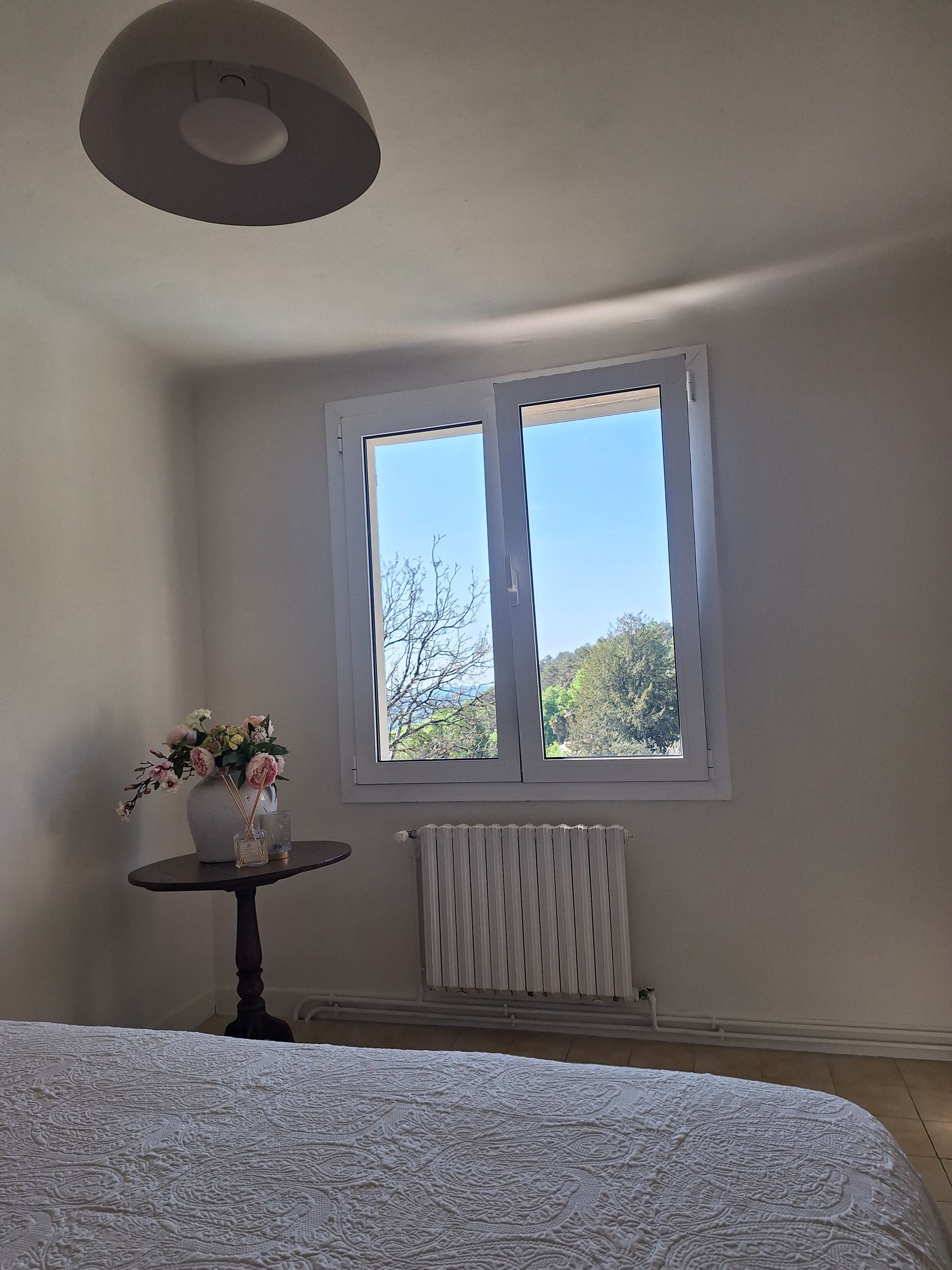 Ein Schlafzimmer mit einem großen Bett, einem kleinen Tisch mit Blumenvase, einem Heizkörper und einem Fenster mit Blick auf Bäume.