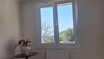 Ein Schlafzimmer mit einem großen Bett, einem kleinen Tisch mit Blumenvase, einem Heizkörper und einem Fenster mit Blick auf Bäume.