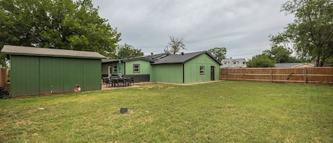A green house with a fenced backyard, a patio table, and chairs.