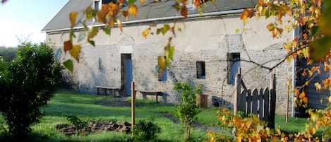 A rustic stone building with a sloped roof, a small garden with grass and a few trees, and a wooden fence.