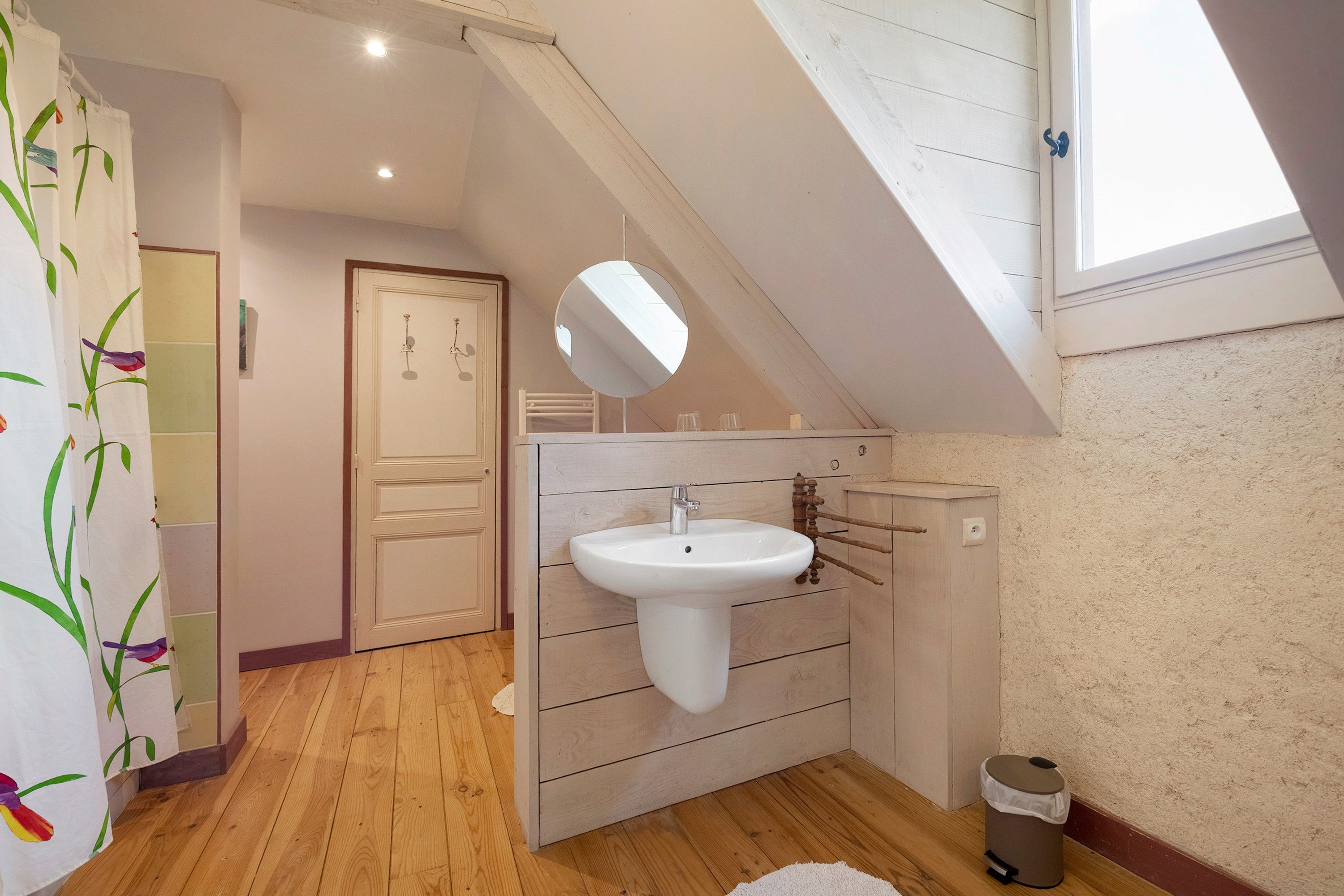 Une salle de bain avec un plancher en bois, un lavabo blanc, un miroir et une porte donnant sur une autre pièce.