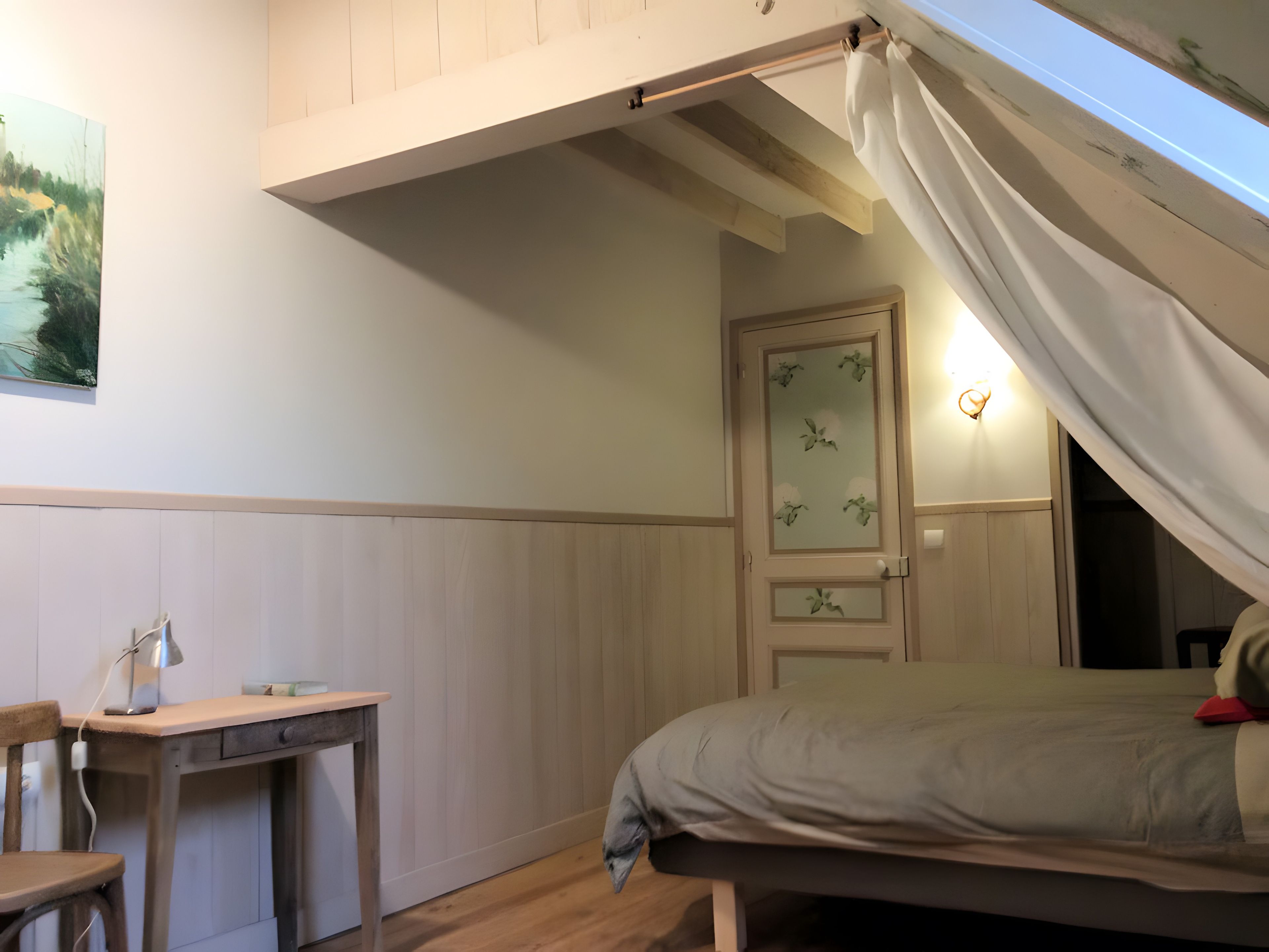 A bedroom with a bed, a desk, and a chair. There is a door with a glass panel and a light fixture above it.