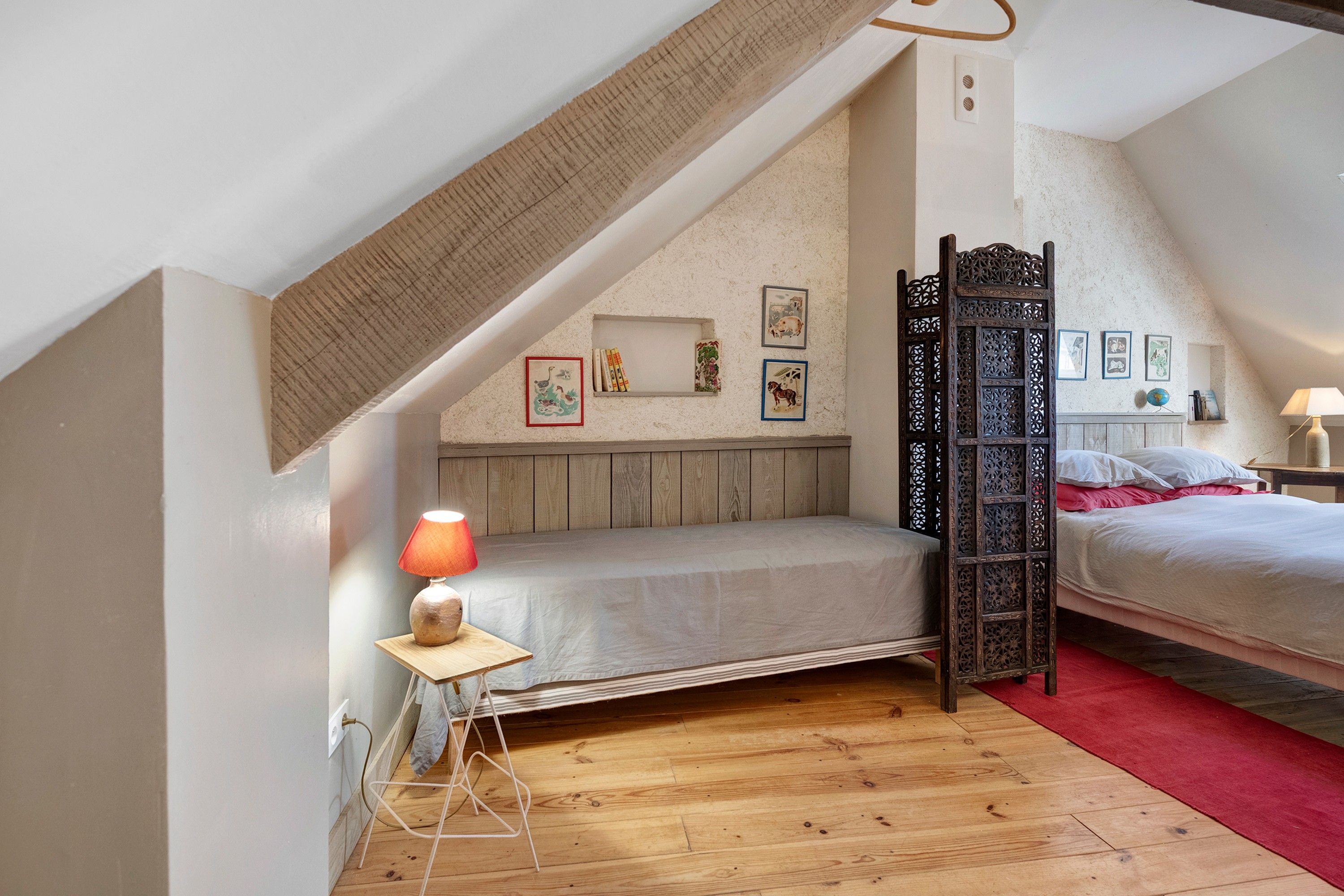 Ein Schlafzimmer mit schrägem Dach, Holzboden, zwei Betten, einem Nachttisch mit Lampe und Wanddekorationen.