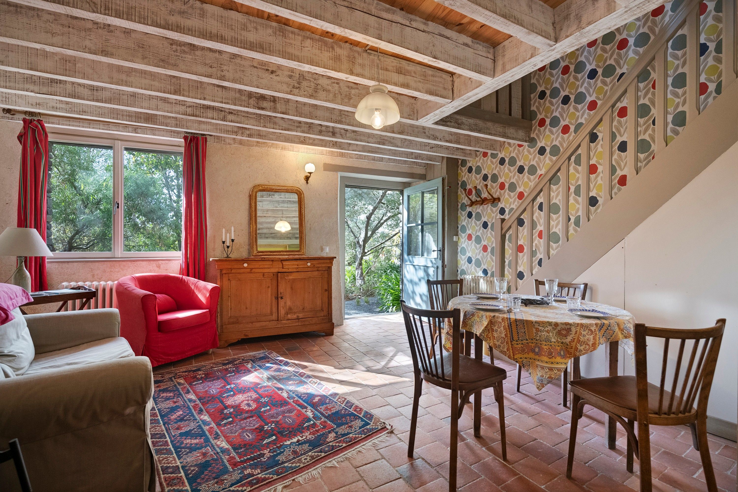 A room with a wooden ceiling, a dining table set with chairs, a red armchair, a cabinet, and a patterned rug.