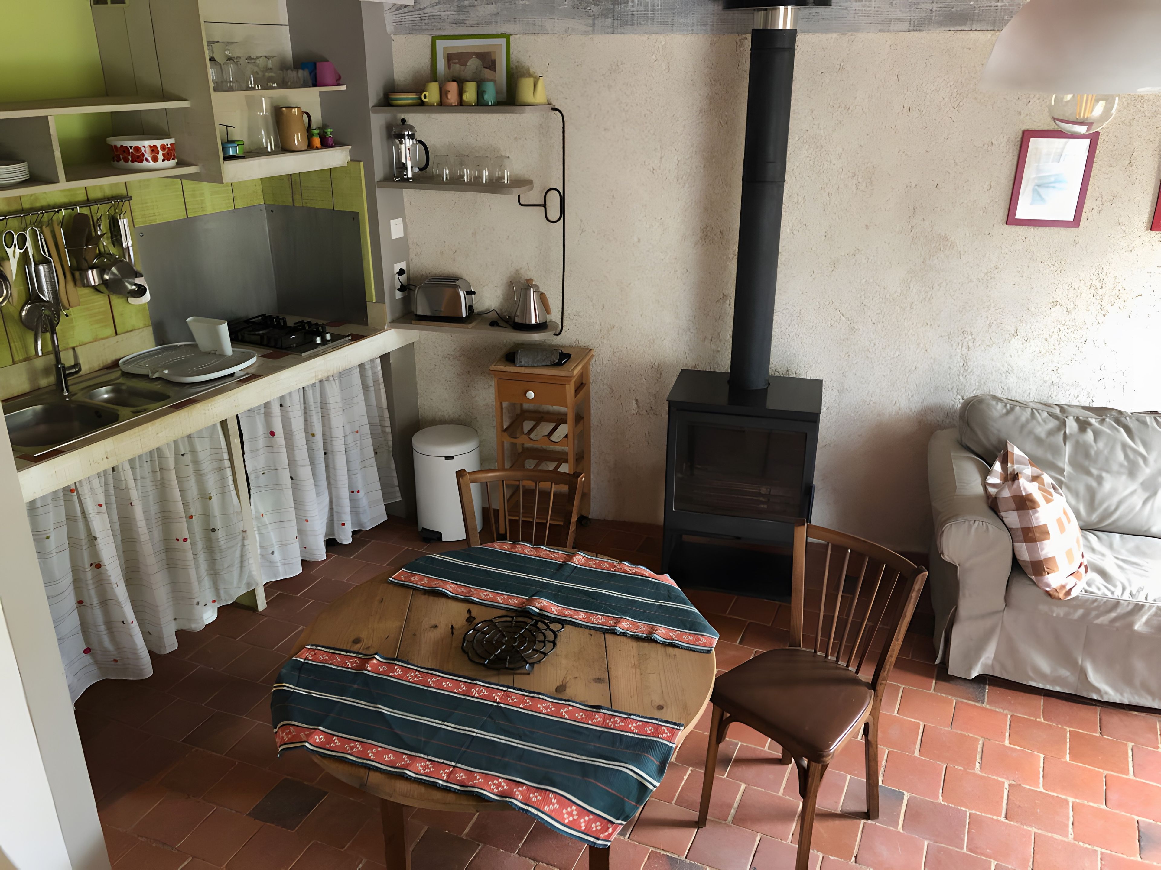 A cozy living space with a kitchen area, a dining table with a patterned tablecloth, a sofa with a checkered pillow, and a wood-burning stove.
