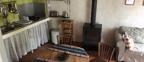 A cozy living space with a kitchen area, a dining table with a patterned tablecloth, a sofa with a checkered pillow, and a wood-burning stove.