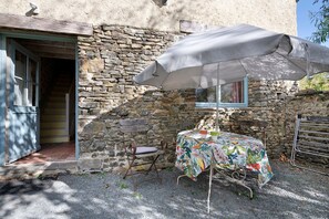 A rustic stone building with a wooden door, a small table with a floral tablecloth, and a metal chair. - BiâO Petitgîte holiday home with terrace, shared garden, and Wi-Fi (Bretteville-sur-Ay)