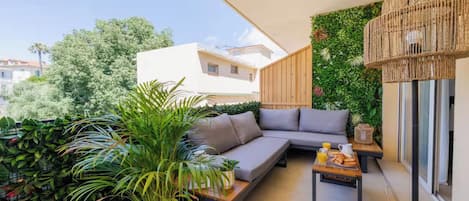 A balcony with a grey sofa, a small table with a tray of food, and lush greenery.