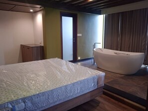 A bedroom with a mattress, a wooden cabinet, a bathtub, and a door leading to another room. - Soul by Araiya, Paithalmala, A Vihaara Resort & Spa (Kannur)