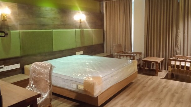 A hotel room with a wooden bed, a wooden desk, a chair, a small table, and a wall-mounted lamp.