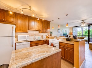 A modern kitchen with wooden cabinets, granite countertops, a built-in oven, and a large island with a sink. - Kona Pacific C421 | Walk to Aliʻi Drive & Beach (Kailua-Kona)