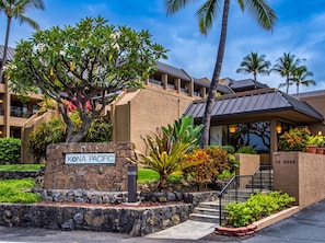 A hotel entrance with a stone sign, a palm tree, and a building with a sloped roof. - Kona Pacific C421 | Walk to Aliʻi Drive & Beach (Kailua-Kona)