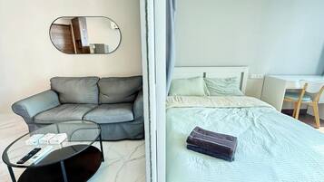 A hotel room with a sofa, a round table with a remote control, a bed with a pillow and a blanket, and a desk with a chair.