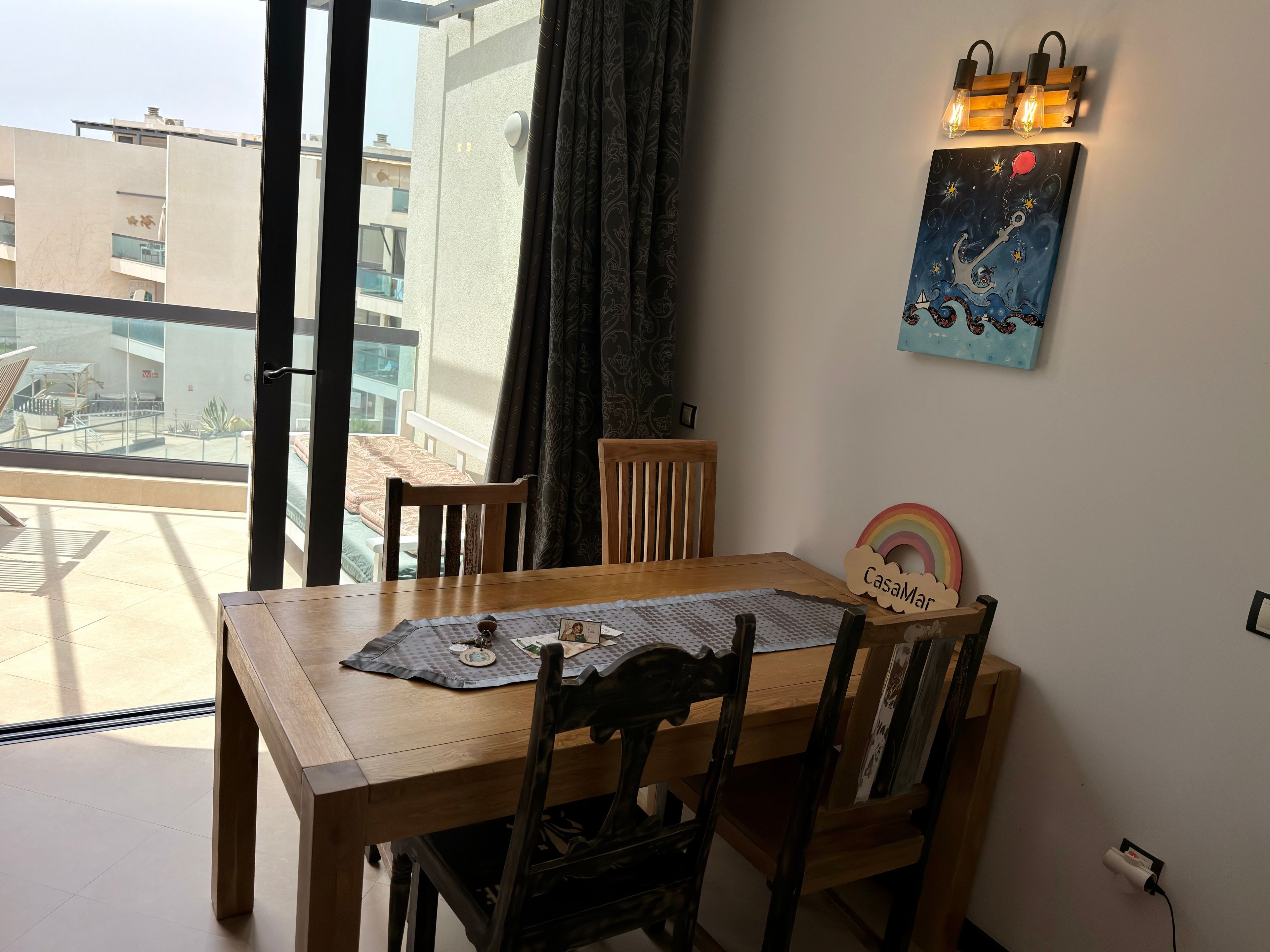 A dining area with a wooden table and chairs, a placemat, a picture frame, and a wall-mounted light fixture.