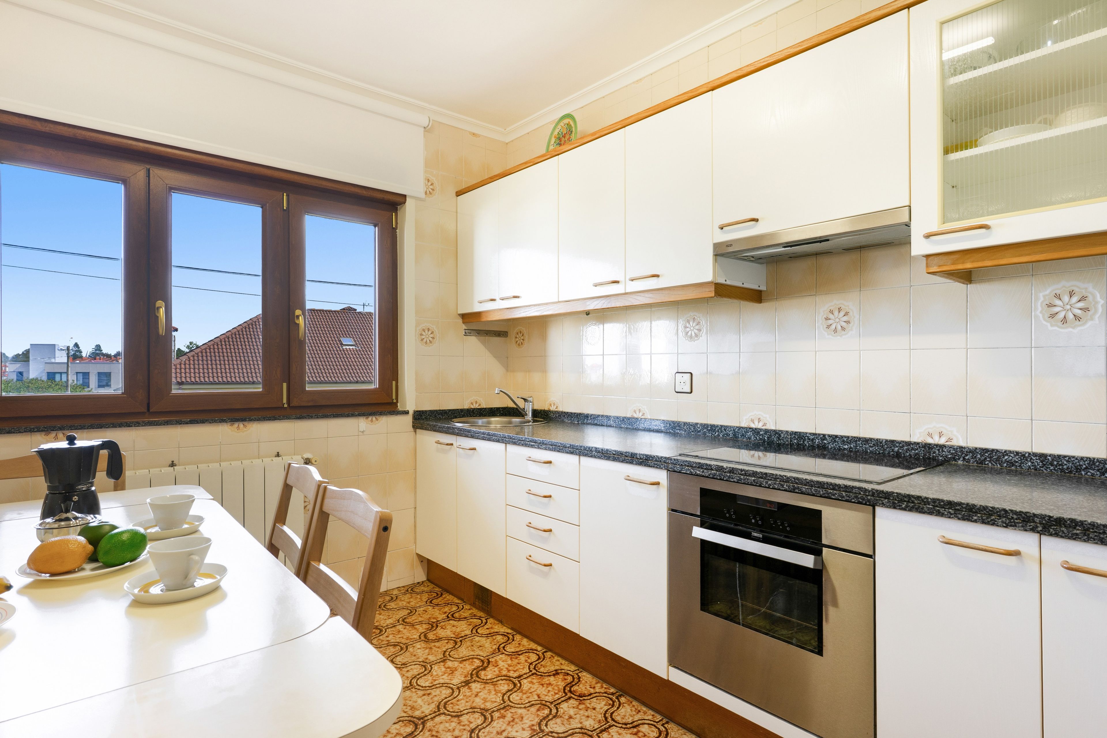 Una cocina con armarios blancos, horno de acero inoxidable y ventana con vista a edificios.