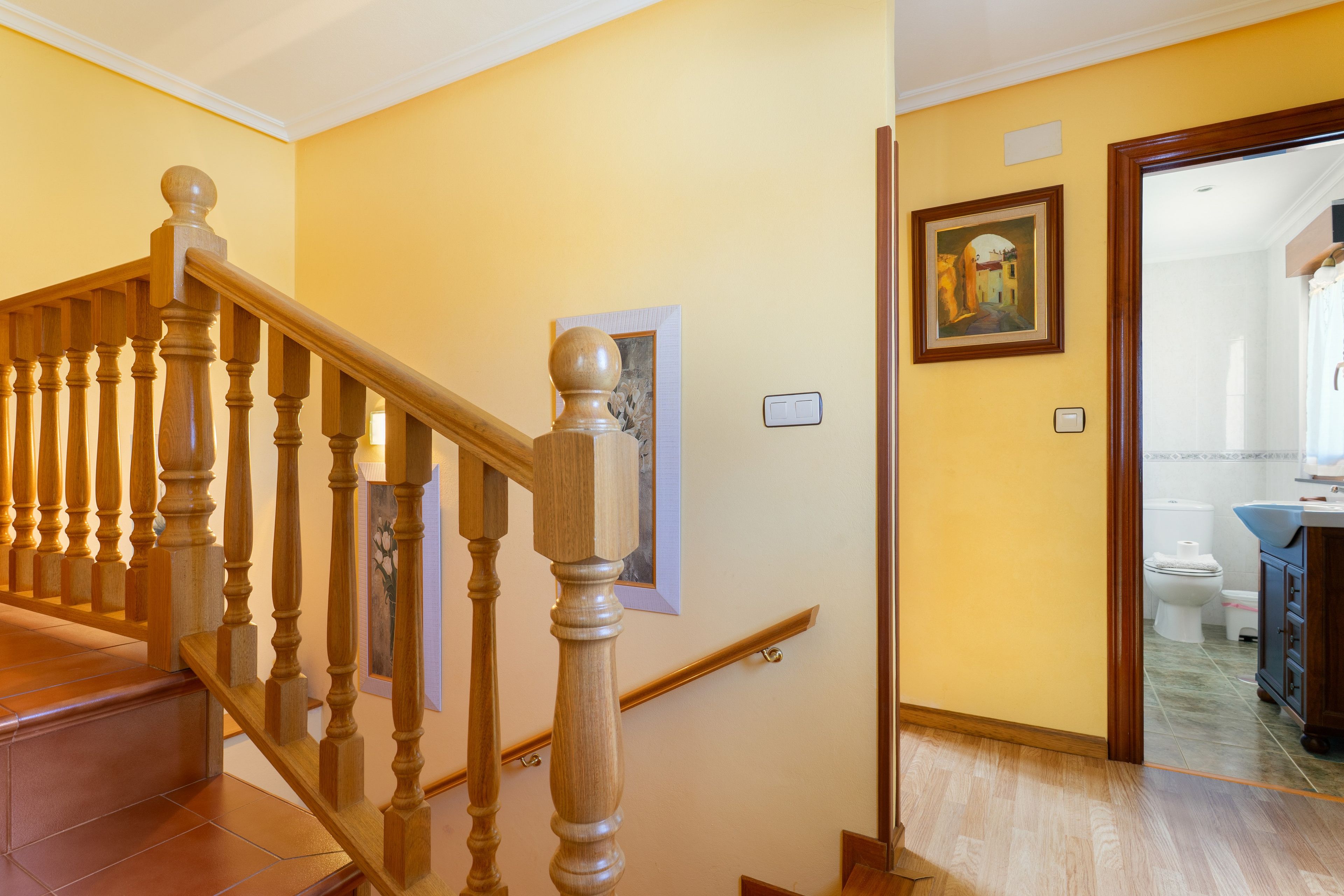 Una escalera de madera con barandilla, un cuadro en la pared y un baño visible a través de una puerta abierta.