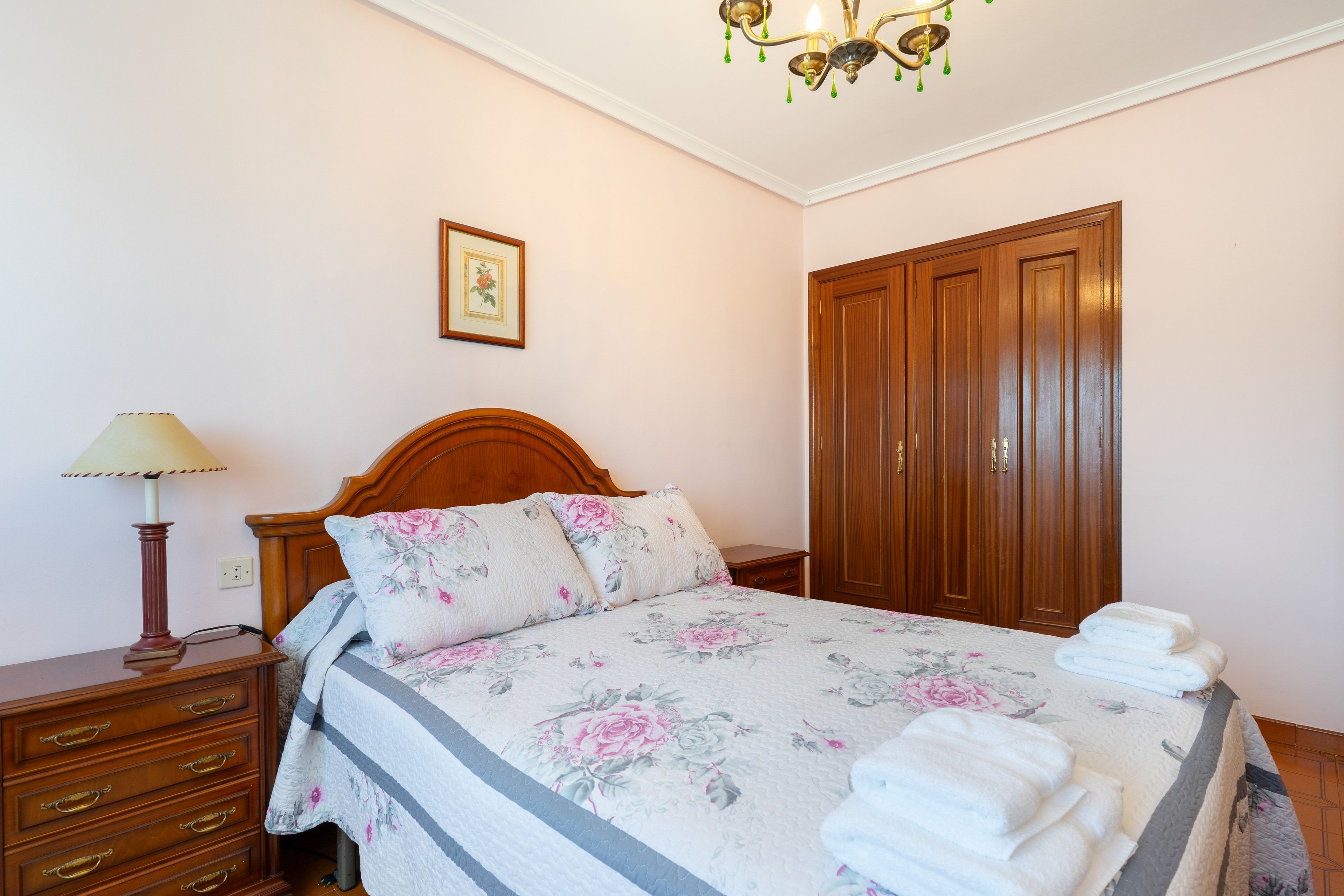 Een slaapkamer met een houten bed, een nachtkastje, een lamp, een kledingkast en een ingelijst schilderij aan de muur.