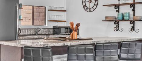 A modern kitchen with a bar area, featuring a marble countertop, black bar stools, and a decorative clock.