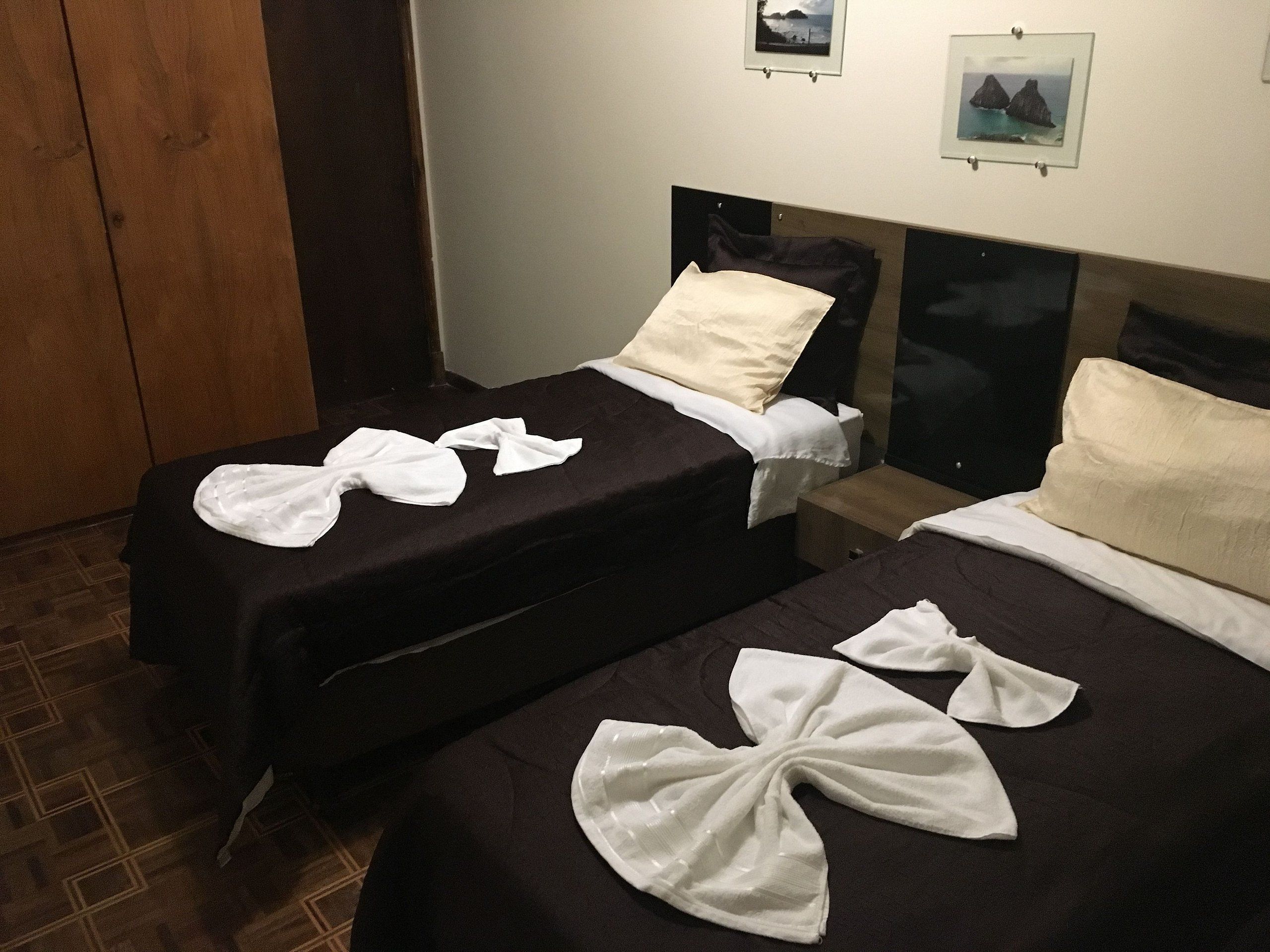 Two single beds with white towels folded on them, a wooden wardrobe, and a wall with framed pictures.