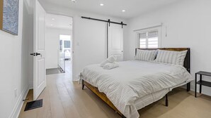 Een slaapkamer met een bed, nachtkastje, raam en deur naar een andere kamer.