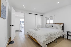 Ein Schlafzimmer mit einem Bett, einem Nachttisch, einem Fenster und einer Tür zu einem weiteren Raum.