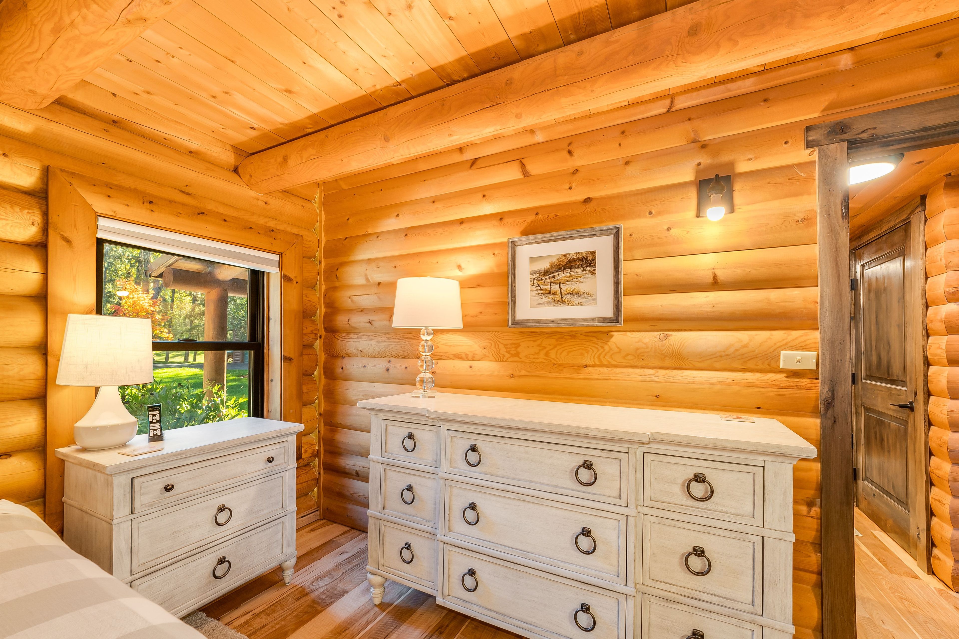Een blokhutkamer met houten plafond, een raam met uitzicht op groen, een dressoir met meerdere lades en een deur.