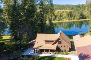 A cabin with a sloped roof, a porch, and a view of a lake surrounded by trees. - Riverfront Cabin on the Swan | Fire Pit & Fishing (Bigfork)
