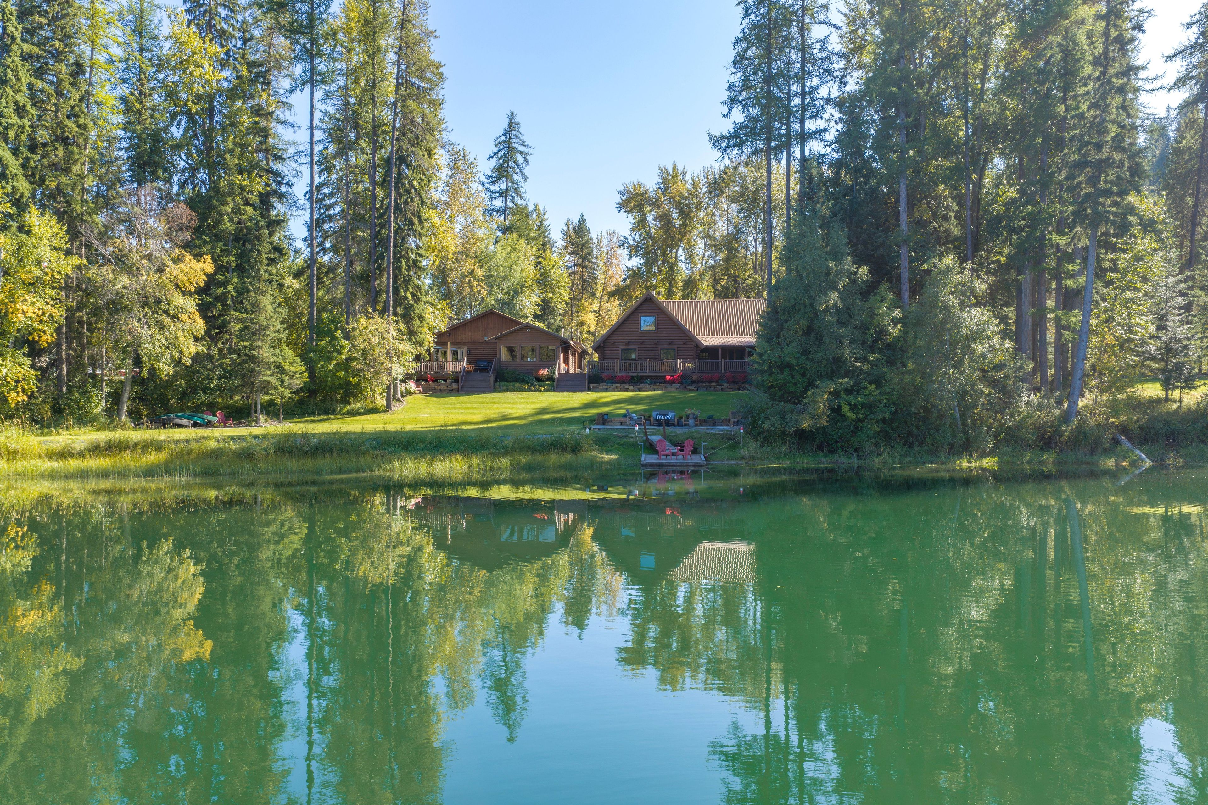 Een lodge met een groot terras en een aanlegsteiger aan een rustig meer, omringd door hoge bomen.
