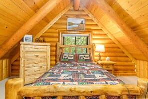Ein Schlafzimmer in einer Blockhütte mit einem Bett, einem Schminktisch und einem Fenster mit Blick auf Bäume.