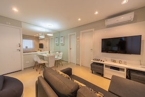 Una sala de estar moderna con un televisor de pantalla plana, sofás grises, una zona de comedor con una mesa y sillas blancas, y una cocina al fondo.