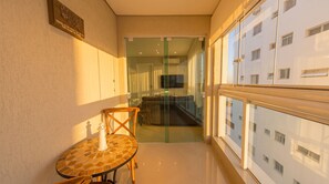 Un estrecho pasillo con una puerta de cristal que da a un comedor, una pequeña mesa con una pieza de ajedrez, y vista a un edificio con múltiples ventanas.