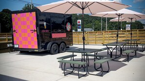 En madvogn med et rosa og orange ternet mønster parkeret ved siden af picnicborde under store parasoller.