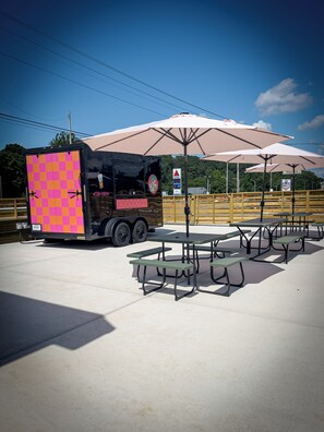 Food truck com design quadriculado em rosa e laranja estacionado ao lado de mesas de piquenique sob grandes guarda-sóis.
