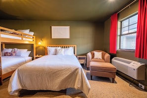 Quarto de hotel com beliche, uma cama de solteiro, uma cadeira, aquecedor e janela com cortinas.