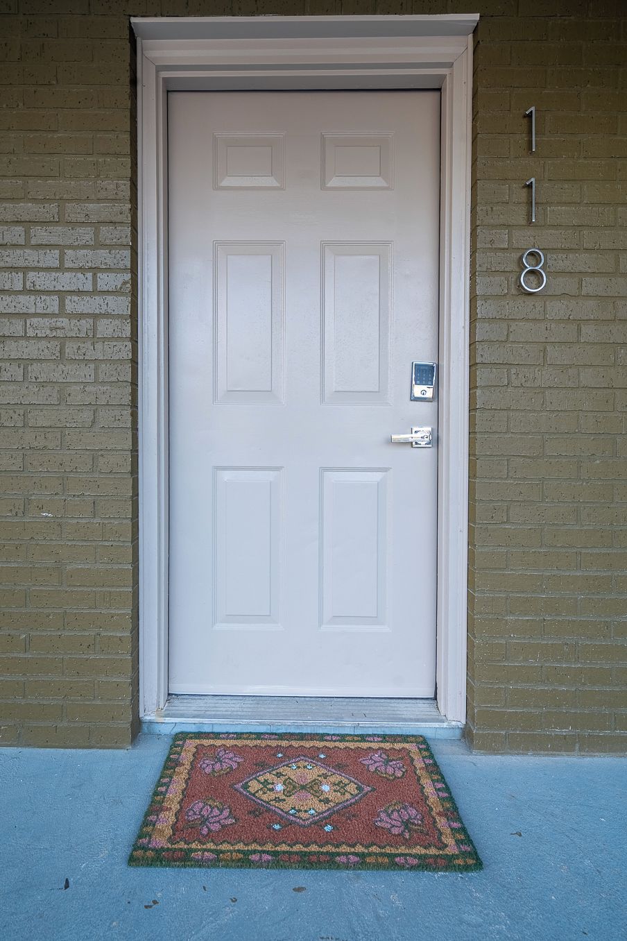 A white door with a keypad lock, a doormat, and a brick wall with the number 18 visible.