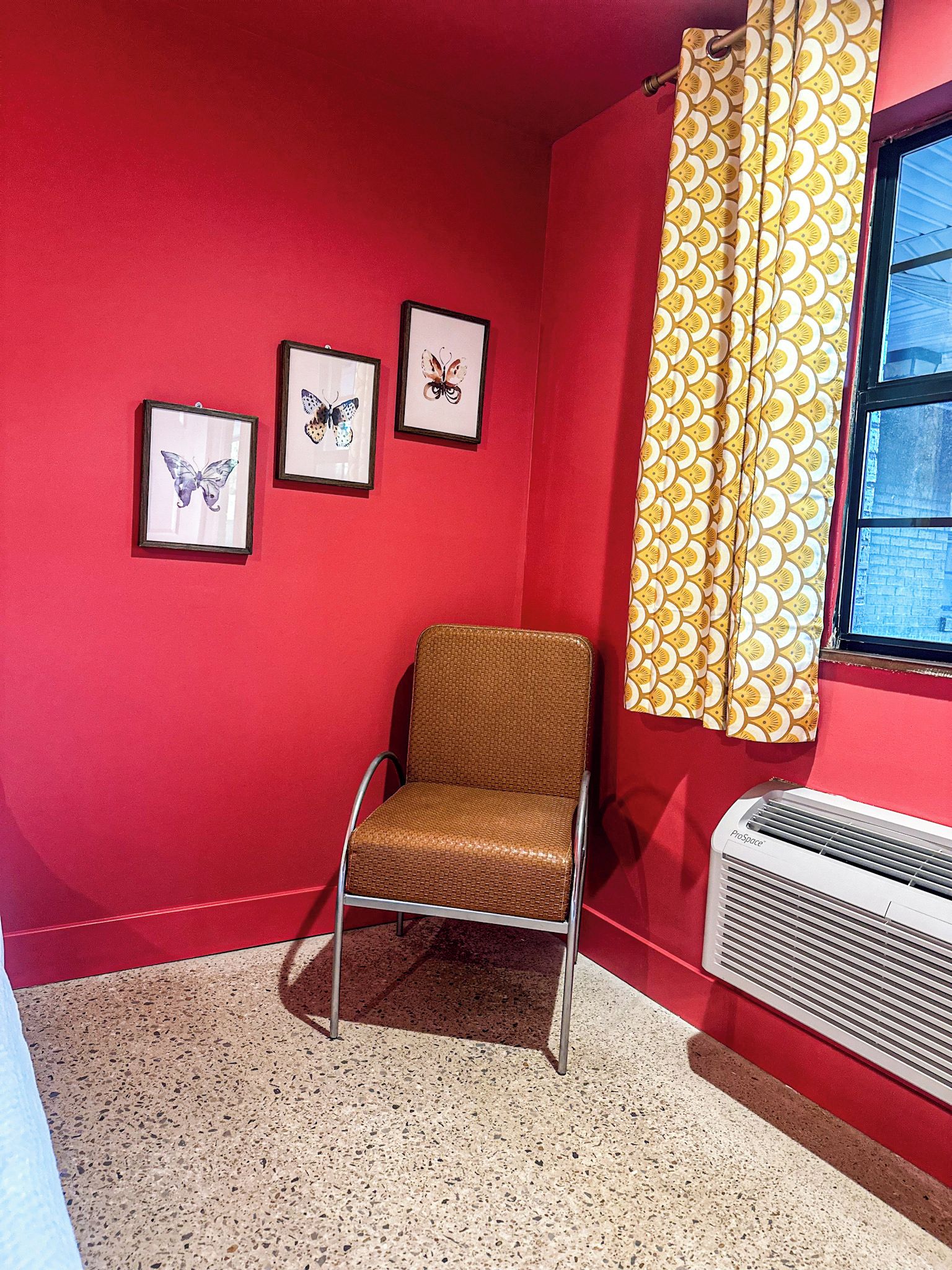A room with a red wall, a brown chair, a window with yellow curtains, and a ProSpace air conditioner.